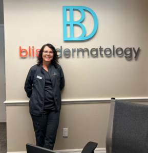 Employee Angela Sommers smiles in front of the Bliss Dermatology office reception wall